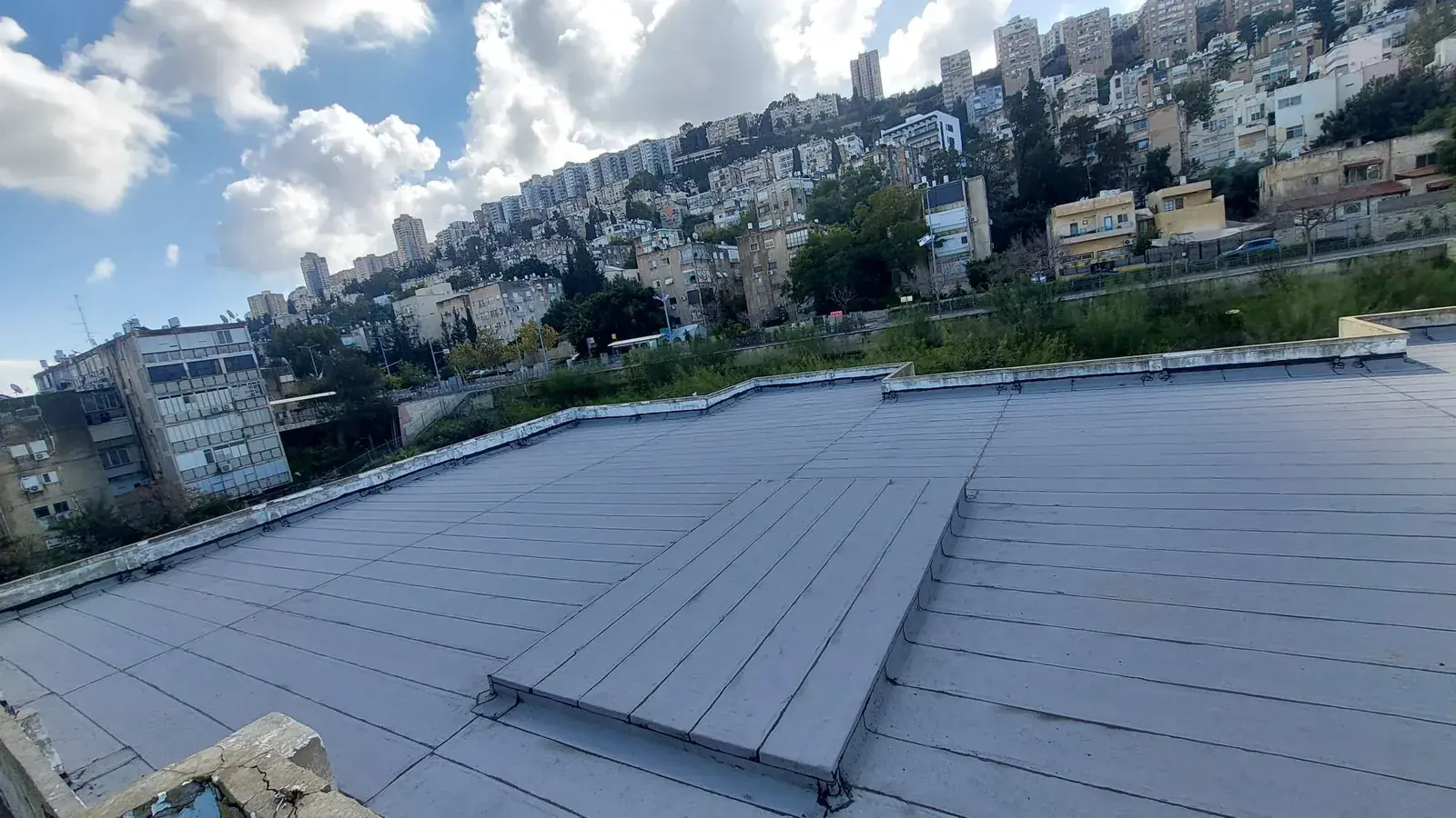 גג שטוח גדול מכוסה ביריעות איטום אפורות בשורות חופפות, עם שכונת מגורים על רכס ברקע